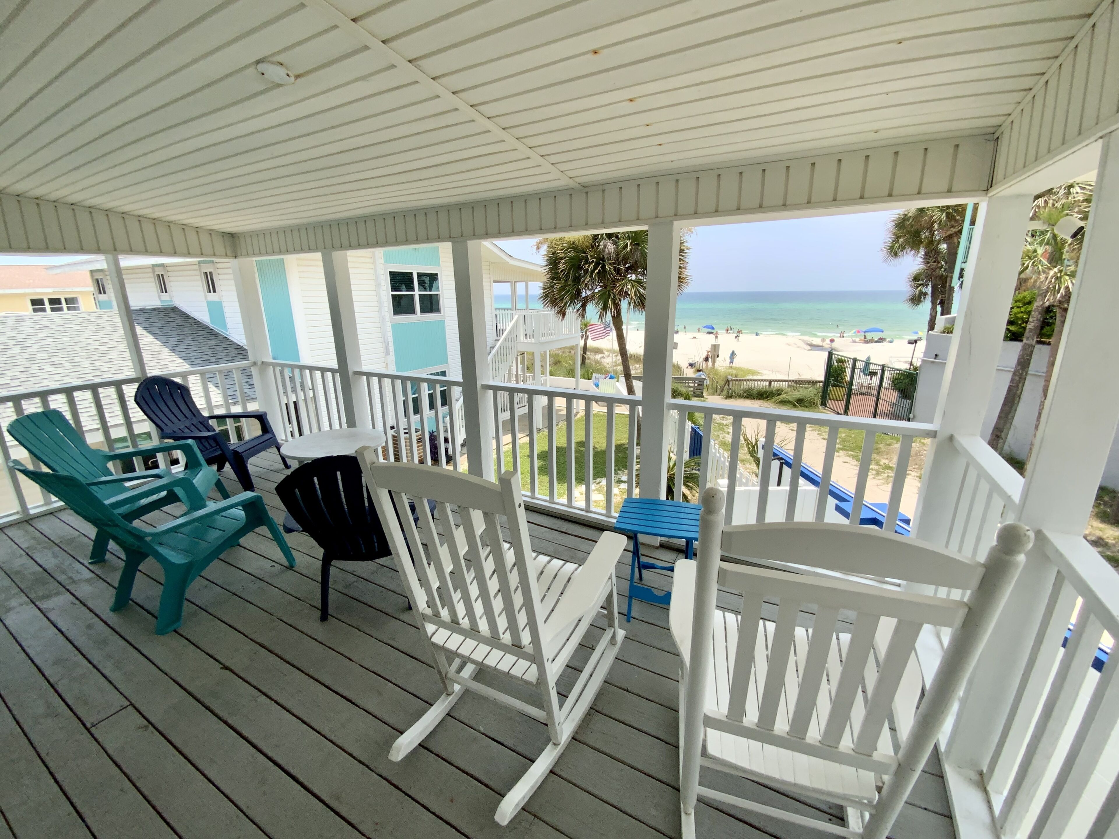 A covered porch with white railings, blue and white Adirondack chairs, and a view of the beach.