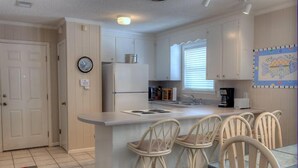 A compact kitchen with white cabinets, a refrigerator, a coffee maker, and a small island with bar stools. - BEACHFRONT PRIVATE COTTAGE, FULL FLOOR, DECK, FREE PARKING BY ENTRANCE, 2BD 2BA  (Panama City Beach)