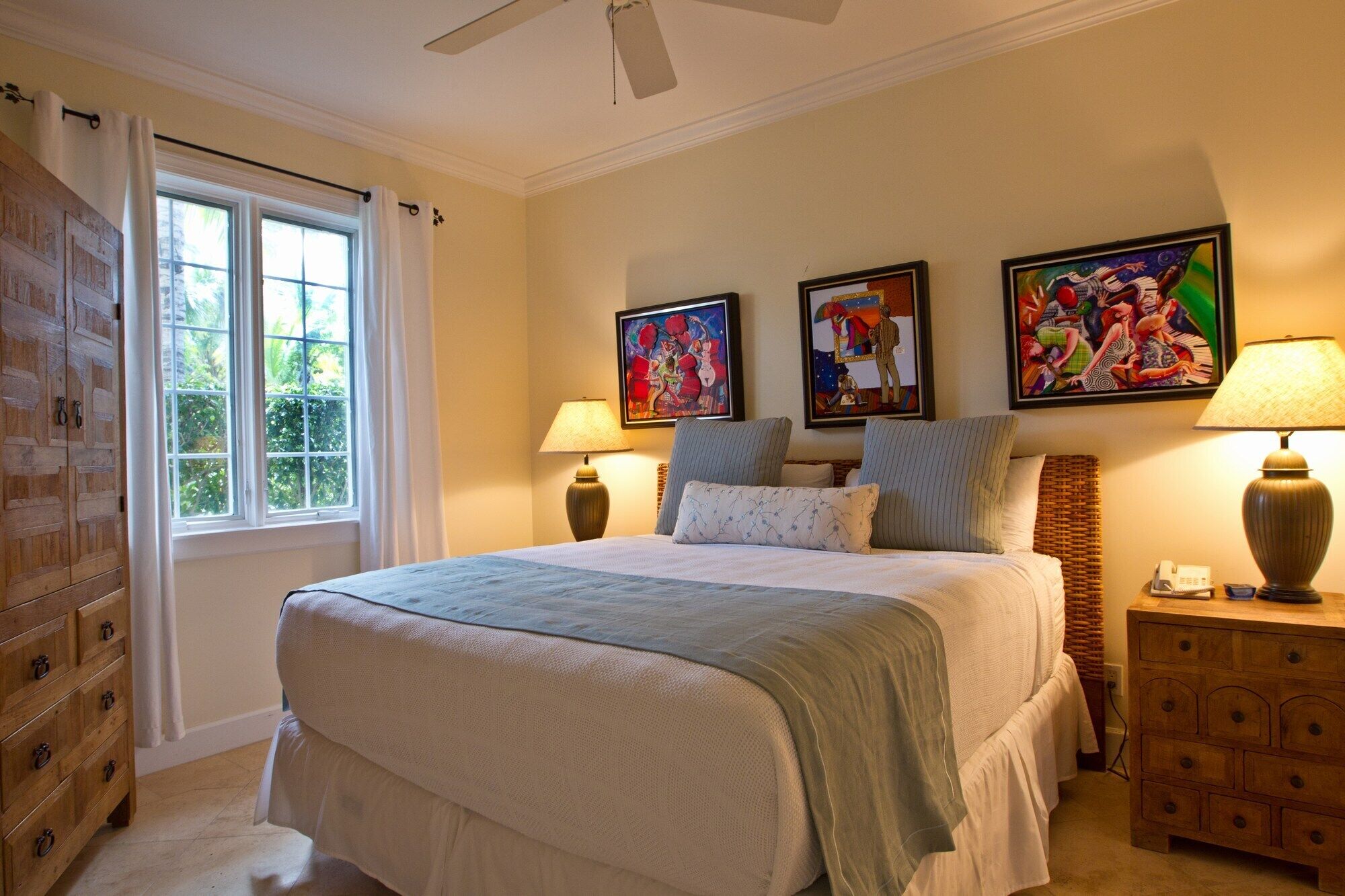 A bedroom with a large bed, two bedside lamps, a wooden dresser, and three framed artworks on the wall.