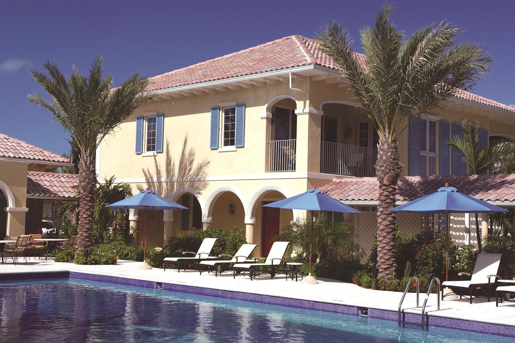 A two-story hotel with a swimming pool, palm trees, and blue umbrellas.