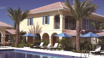 A two-story hotel with a swimming pool, palm trees, and blue umbrellas.