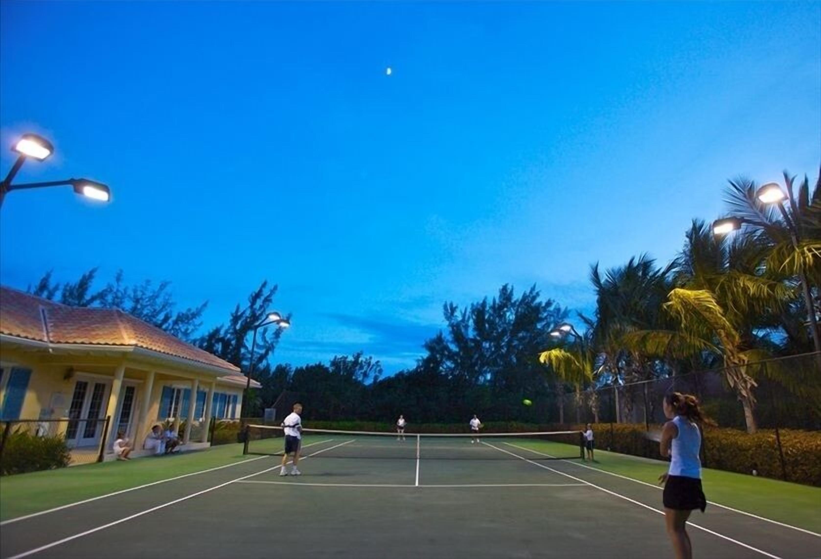 An outdoor tennis court at dusk with a house on one side and palm trees on the other.