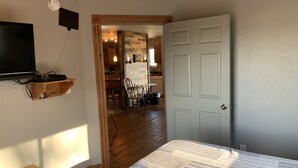 A bedroom with a bed, a TV mounted on a wooden shelf, a door, and a view into the dining area.