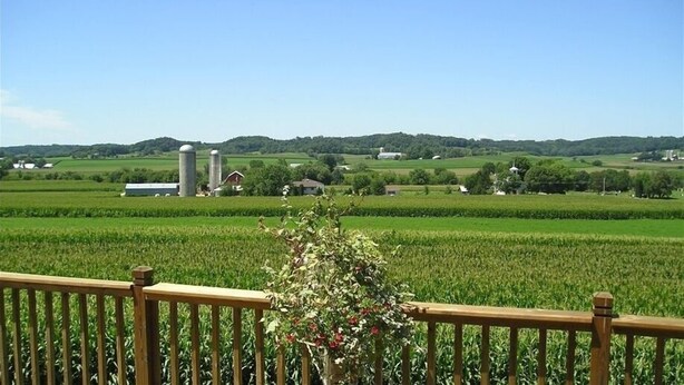 A rural landscape with a wooden fence, a field of crops, and distant buildings including a silo and a house.