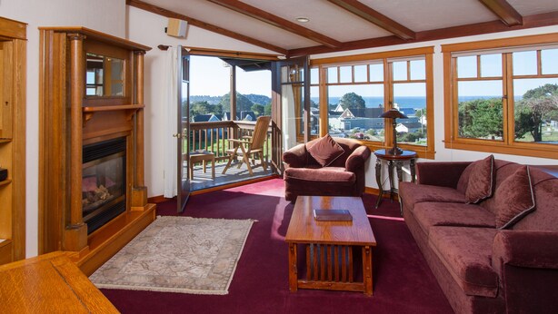 A cozy living room with a fireplace, a red couch, a wooden coffee table, and large windows offering a view of the outdoors.