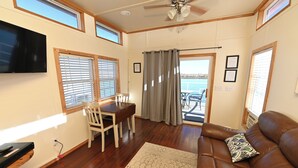 A living room with a brown leather sofa, a dining table with chairs, a TV mounted on the wall, and a view of the water through the windows. - Rebel's Rest - A Romantic Getaway Houseboat w/ New Water Filtration System (Apalachicola)
