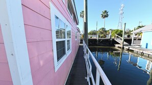 A pink building with a dock extending into calm water, surrounded by palm trees and a clear sky. - Rebel's Rest - A Romantic Getaway Houseboat w/ New Water Filtration System (Apalachicola)