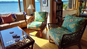 Una sala de estar acogedora con un sofá, dos sillones, una mesita y una estantería. Amplias ventanas ofrecen vista al mar.