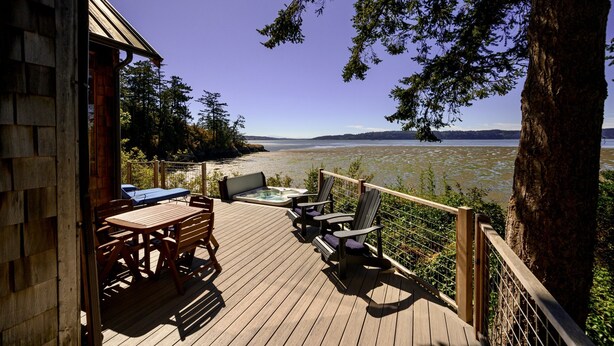 Una terraza de madera con mobiliario exterior, un jacuzzi y vistas a un lago y montañas lejanas.