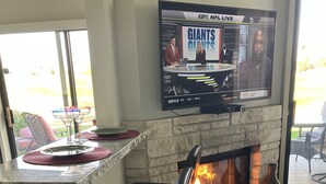 Una cocina moderna con televisión integrada mostrando un canal deportivo, chimenea de piedra y zona de bar con taburetes.