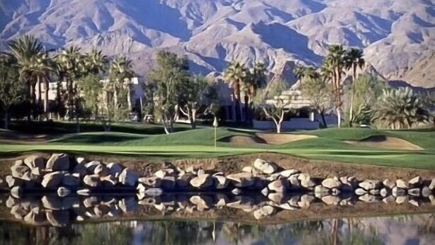 Un campo de golf con un estanque que refleja el paisaje, palmeras y una montaña al fondo.