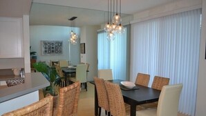 Un comedor moderno con una mesa de madera oscura y sillas de mimbre, un área de cocina con armarios blancos y un amplio ventanal con cortinas traslúcidas.
