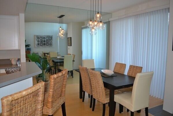 Un comedor moderno con una mesa de madera oscura y sillas de mimbre, un área de cocina con armarios blancos y un amplio ventanal con cortinas traslúcidas.