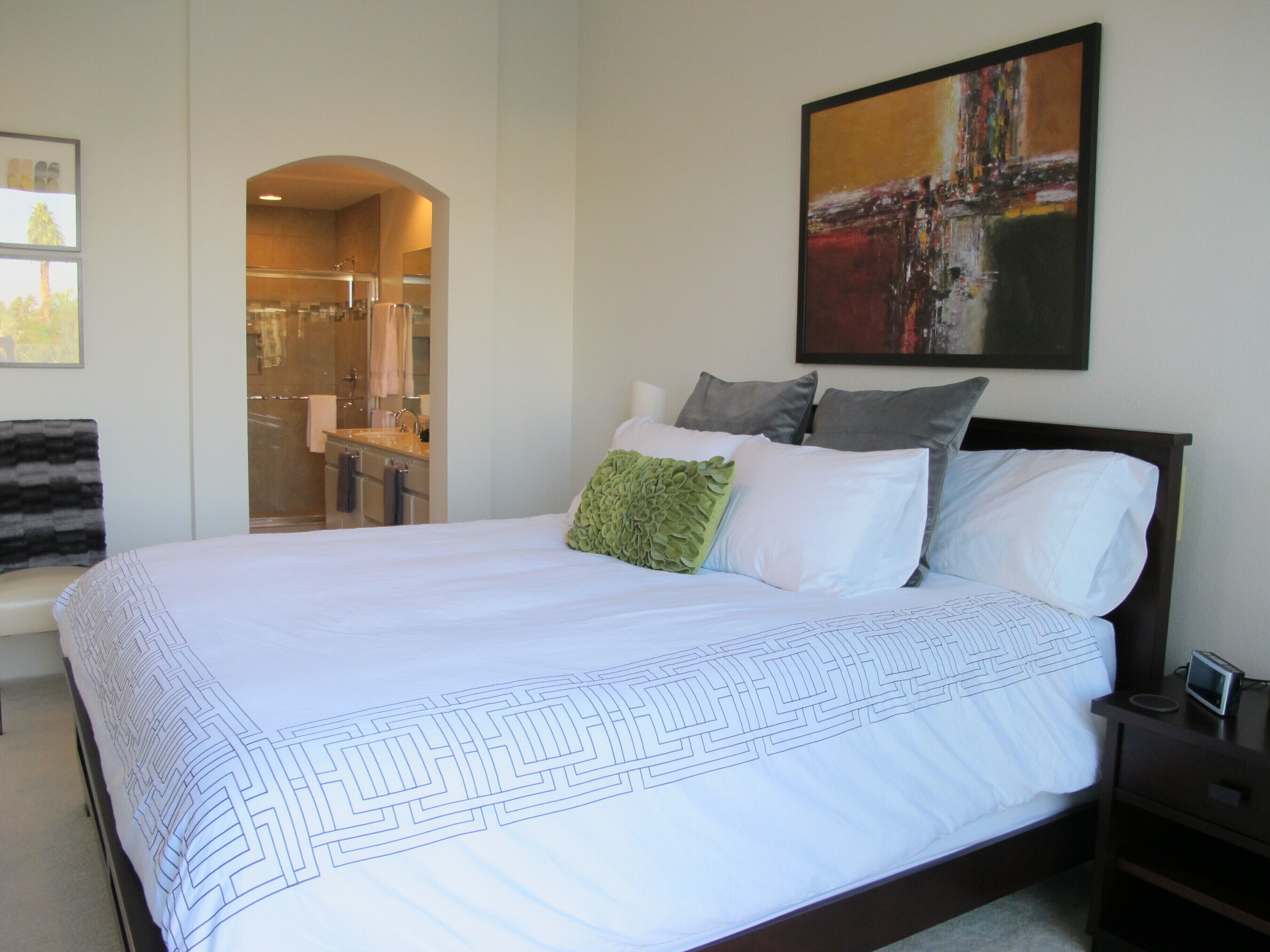 A bedroom with a large bed, a nightstand, a painting on the wall, and a bathroom visible through an archway.