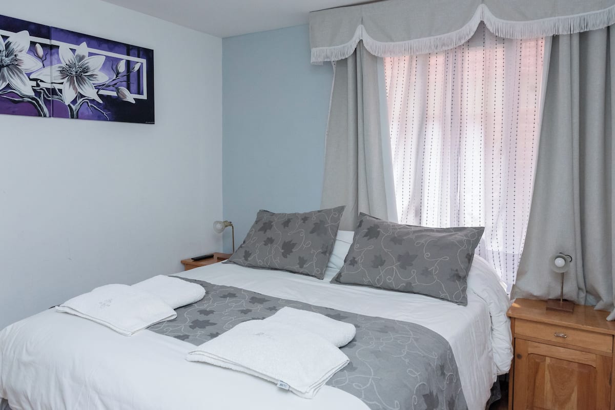 Een net opgemaakt bed met witte en grijze lakens, een nachtkastje met een lamp, en een bloemen schilderij aan de muur.