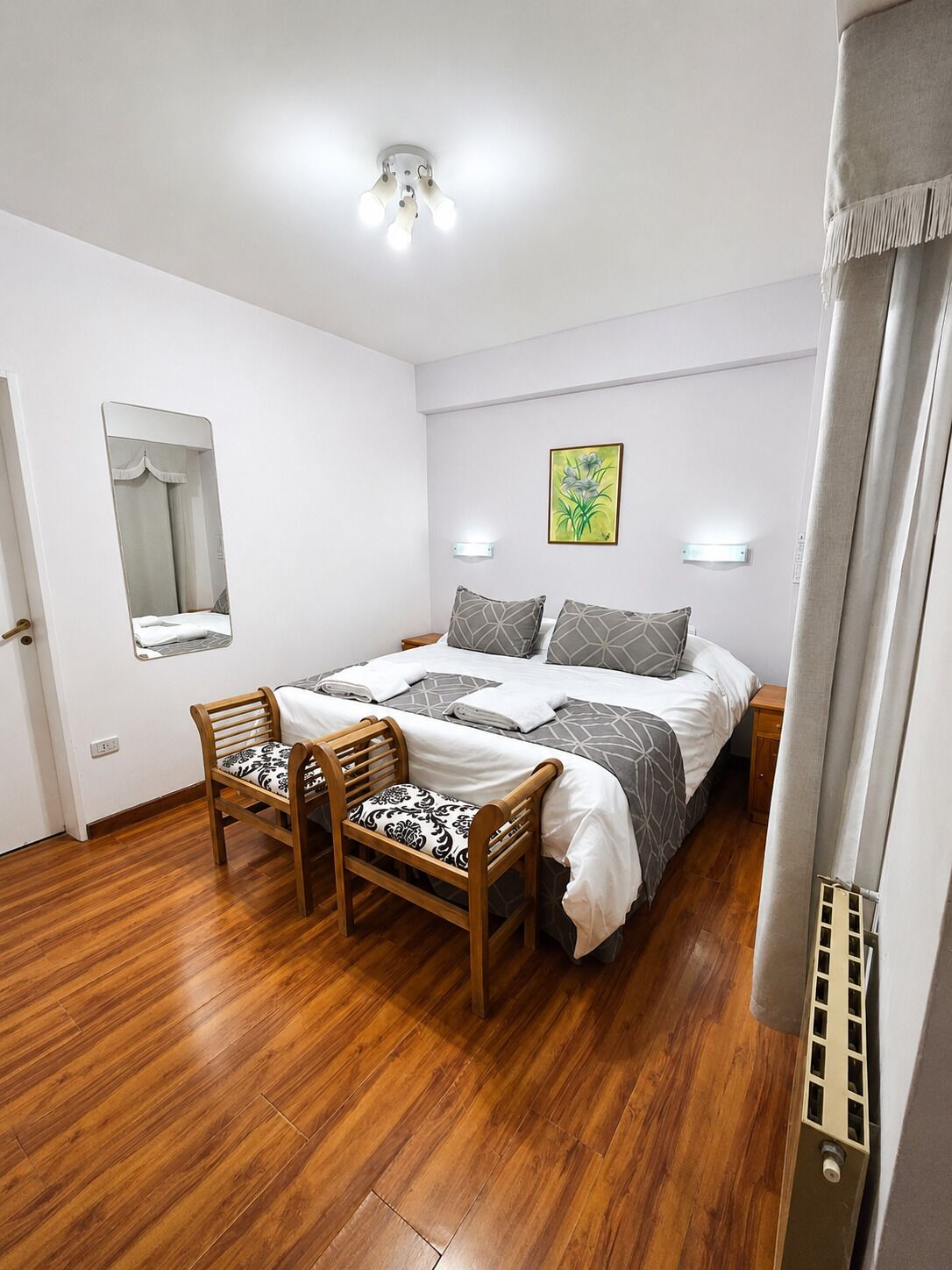 Un dormitorio con cama, dos mesitas de noche, un espejo, una silla y una ventana con cortinas.