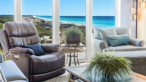 A modern living room with a leather recliner, a sofa, and a small table with a plant. Large windows offer a view of a beach and ocean. - Tops`l Beach Manor 803 - Gorgeous Views! (Miramar Beach)