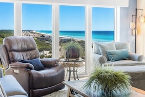 A modern living room with a leather recliner, a sofa, and a small table with a plant. Large windows offer a view of a beach and ocean.