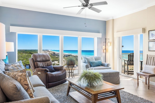 A spacious living room with a view of the ocean, a ceiling fan, and a balcony with a chair.