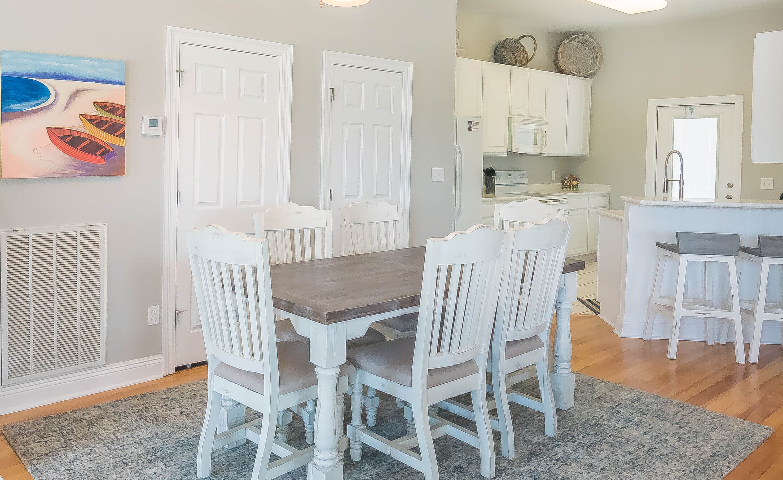 A dining area with a wooden table and white chairs, a kitchen with white cabinets, and a painting on the wall.