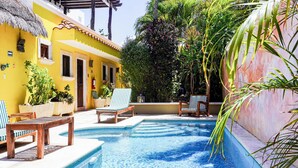 A pool area with lounge chairs and potted plants, surrounded by a yellow building with a tiled roof. - One Bedroom Suite, Serene, Pool, Gardens, Rooftop Terrace with Views (Isla Mujeres)