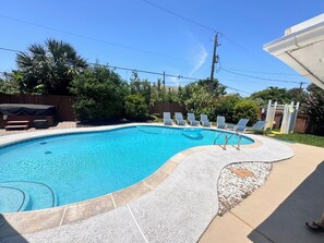 A rectangular swimming pool with a tiled edge, surrounded by a patio area with lounge chairs and a covered seating area.