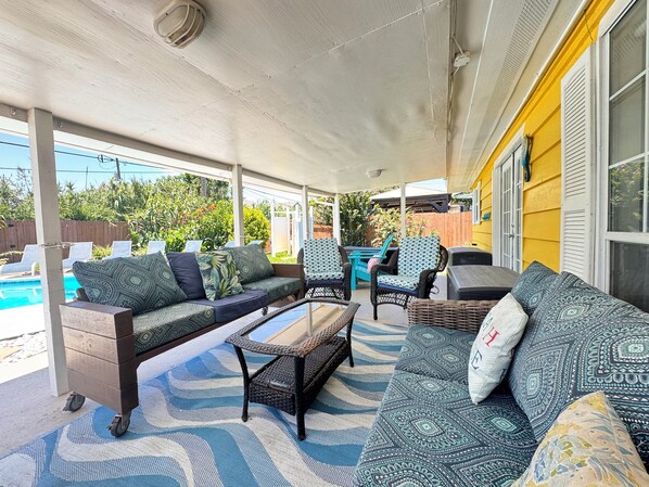A covered patio with a pool, outdoor furniture, and a yellow building in the background.