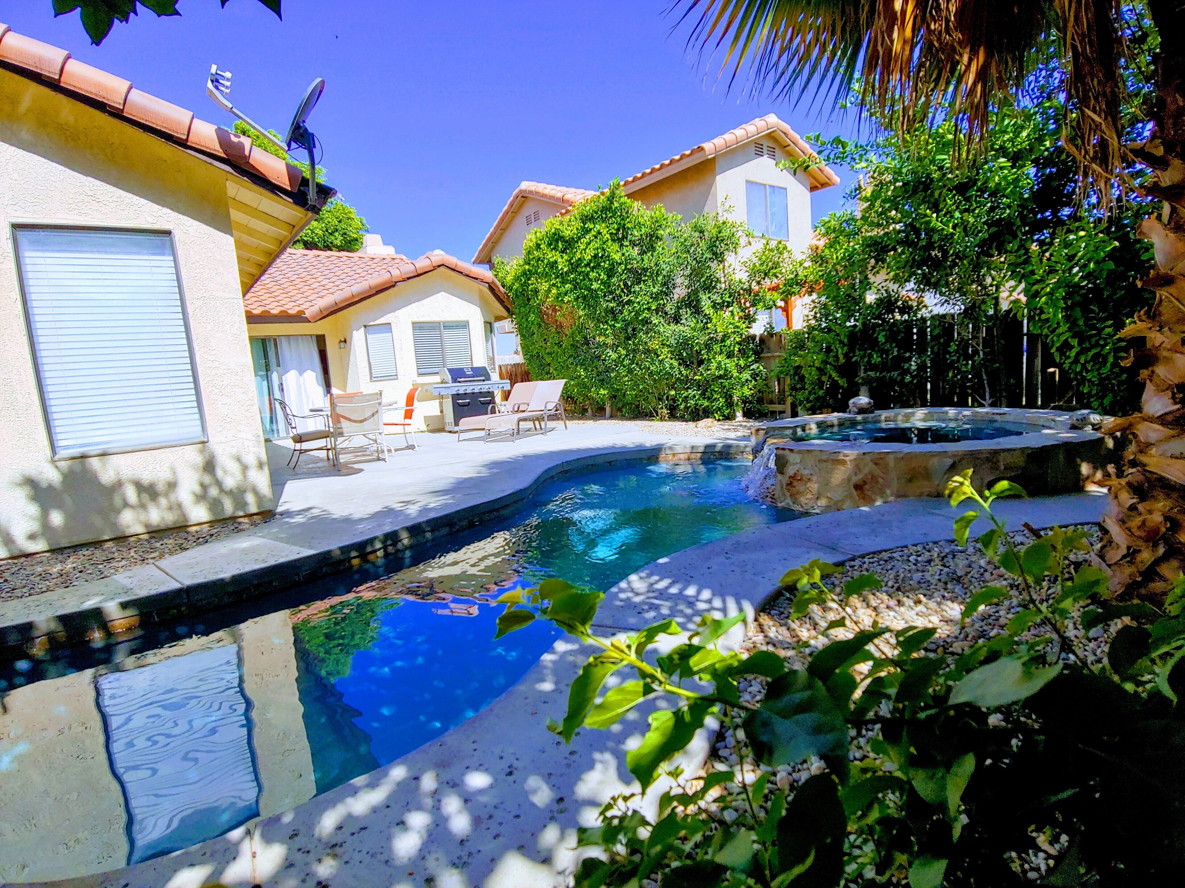 Ein Schwimmbad mit Whirlpool, umgeben von einer Terrasse mit Gartenmöbeln und Blick auf ein Wohngebäude mit Ziegeldach.