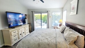 A bedroom with a bed, a television on a dresser, a window with blinds, and a view of the outdoors.