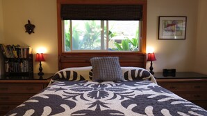 A neatly made bed with a patterned bedspread, a window with a view of greenery, a bookshelf with books, and two bedside lamps.