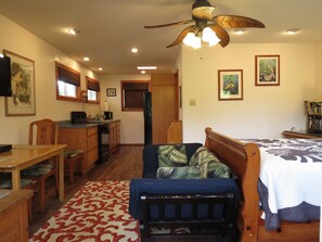 A compact room with a bed, a desk, a chair, a kitchenette, and a ceiling fan. - Cottage-Very Close to Beach-Quiet A/C-Full Kitchen with Range/Oven-Deck-Laundry (Hauula)