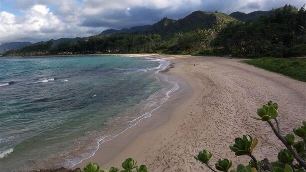 A beach with clear turquoise water, a sandy shore, and lush green hills in the background.