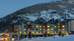 Un hotel de varios pisos con ventanas iluminadas, situado en un paisaje nevado con una montaña al fondo.