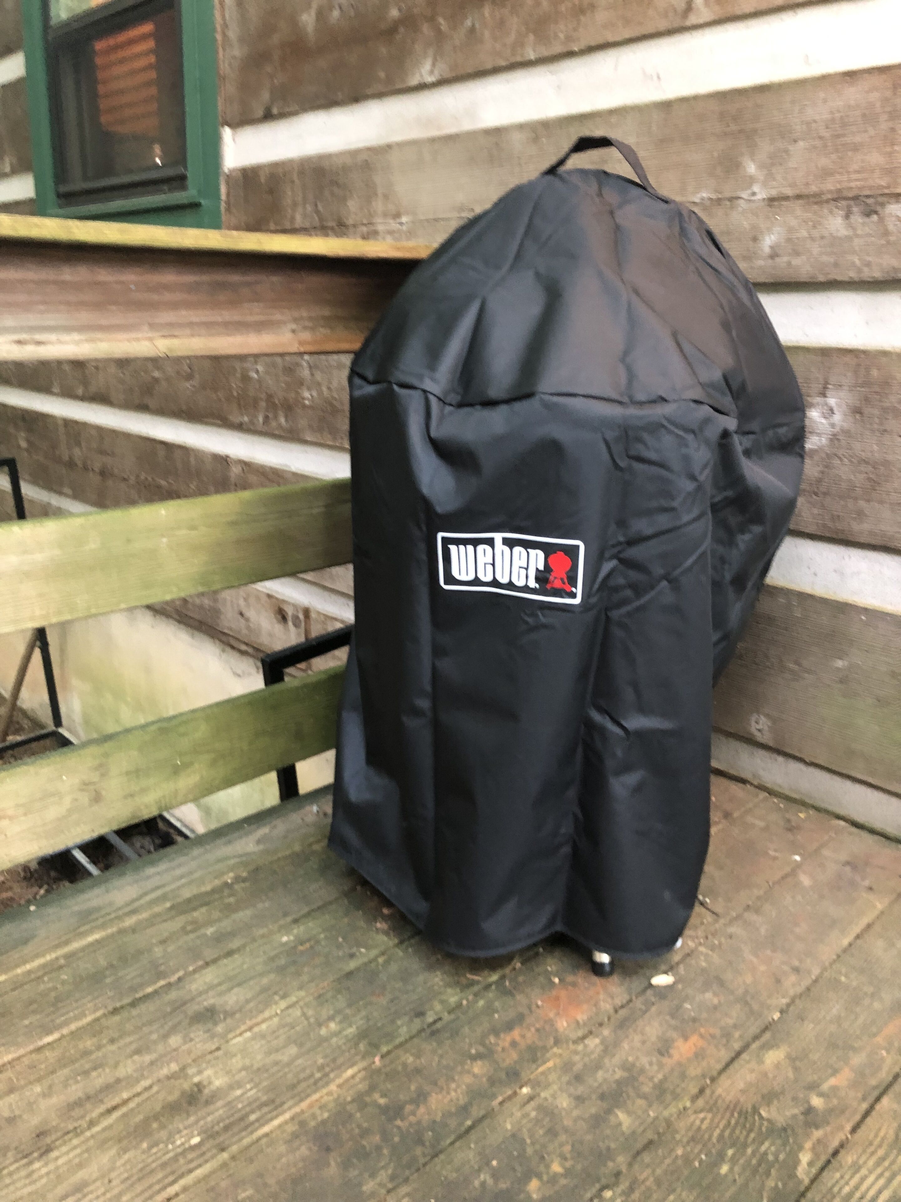 Een zwarte Weber grilltas op een houten bank voor een blokhut.