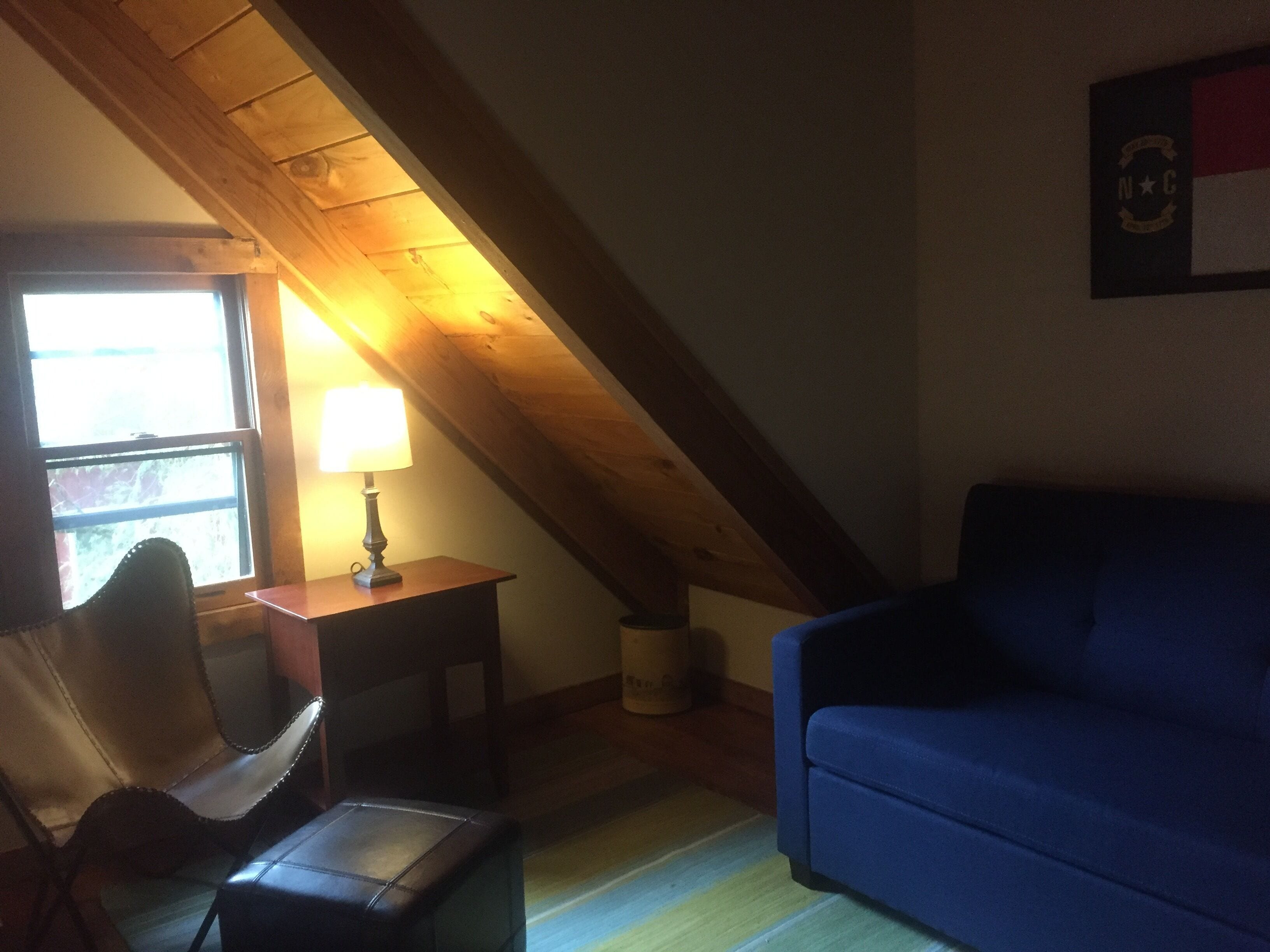 A cozy room with a sloped ceiling, a blue sofa, a small table with a lamp, and a chair.