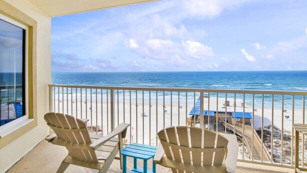 Ein Balkon mit zwei Adirondack-Stühlen und einem kleinen Tisch, mit Blick auf einen Strand mit einer Holzpromenade und einem Gebäude mit blauem Dach.