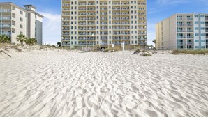 Ein Hotel direkt am Strand mit mehreren Balkonen, ein sandiger Strand im Vordergrund und ein klarer Himmel.
