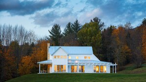 A large, two-story house with a metal roof and multiple windows, surrounded by autumn-colored trees and a grassy hill. - Modern Farmhouse on 25 Acres / Superb Views. (Rochester)