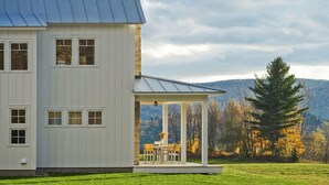A two-story house with a stone chimney, a covered porch with a table and chairs, and a view of a forested hillside. - Modern Farmhouse on 25 Acres / Superb Views. (Rochester)