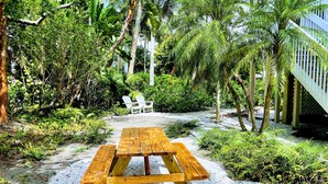 A wooden picnic table surrounded by gravel and greenery, with a white chair and a wooden deck in the background. - Fantastic Island Getaway at Sunset Captiva 48 (Captiva)