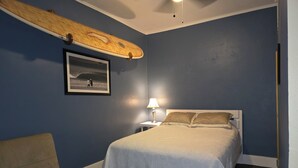 Ein Schlafzimmer mit Bett, Nachttisch, Lampe, einem gerahmten Bild, einer Couch und einem an der Wand befestigten Surfbrett.