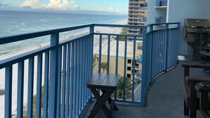 A balcony with a view of the beach and ocean, a barbecue grill, and a wooden table.