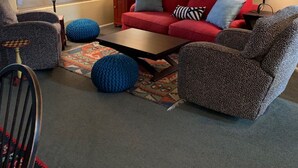 Un salon chaleureux avec un canapé rouge, deux fauteuils à motifs, une table basse et deux lampes. Un tapis coloré et un tableau encadré ornent les murs.
