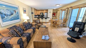 A living room with a sofa, a coffee table, and a black chair. There is a kitchen area with a refrigerator, stove, and bar stools. A painting is on the wall, and a window with curtains is visible. - Spacious Condo w/ FREE WiFi, Parking, Heated Pool, Hot Tubs, Skier Shuttles (Avon)