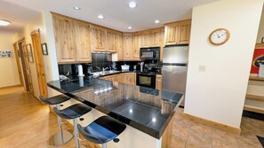 A modern kitchen with wooden cabinets, a stainless steel refrigerator, a microwave, and a large island with bar stools. - Spacious Condo w/ FREE WiFi, Parking, Heated Pool, Hot Tubs, Skier Shuttles (Avon)