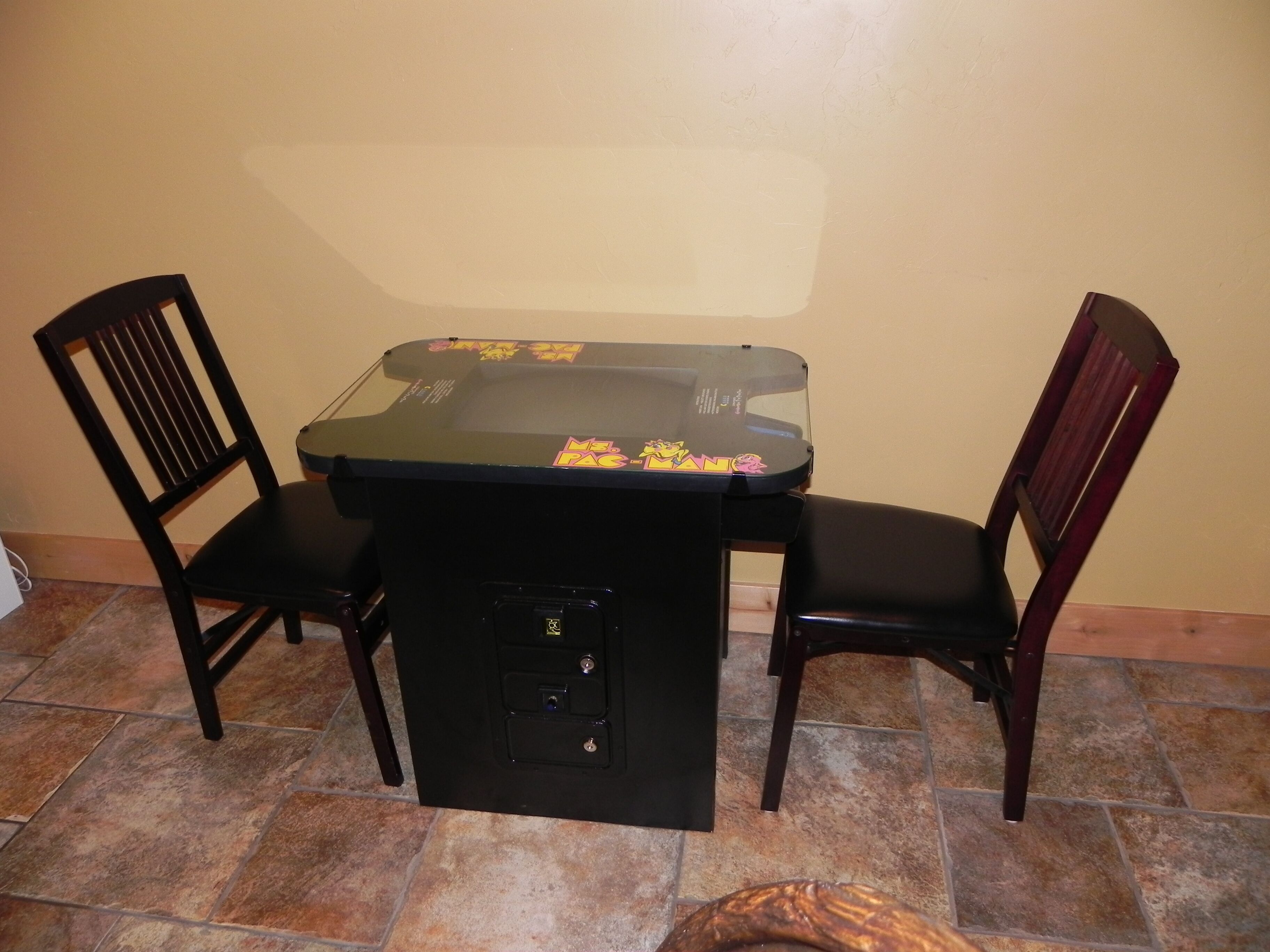 Une petite table de couleur sombre, dotée d’un plateau en verre et ornée de quelques autocollants, entourée de deux chaises en bois de couleur sombre.