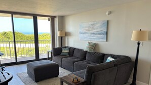 Ein modernes Wohnzimmer mit einem grauen Ecksofa, einem Glastisch und einem kleinen Beistelltisch. Ein großes Fenster bietet einen Ausblick ins Freie.