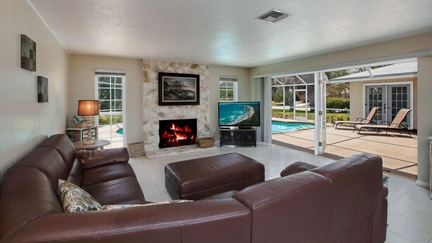A living room with a fireplace, a television, and a pool view. - 5-Min Walk to Beach! Private Heated Pool & 5BR Home (Marco Island)