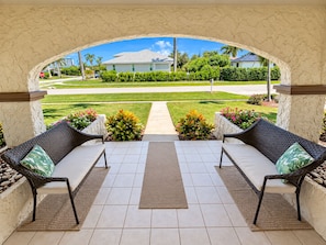 An outdoor patio with two wicker chairs and a central rug, leading to a view of a residential area with a stop sign and greenery.