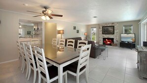 A spacious living area with a dining table, white chairs, a fireplace, a TV, and a stone wall. - 5-Min Walk to Beach! Private Heated Pool & 5BR Home (Marco Island)